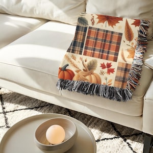 May include: A decorative throw blanket with a patchwork design featuring pumpkins, autumn leaves, and plaid patterns in shades of orange, brown, and cream. The blanket has a fringed edge and is draped over a white sofa.
