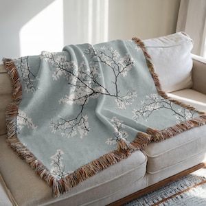 Könnte beinhalten: Hellblaue Decke mit einem floralen Astmuster in Weiß und Schwarz. Die Decke hat einen gefransten Rand in Braun- und Beigetönen. Sie liegt über einem beigen Sofa und sorgt für eine gemütliche Atmosphäre.