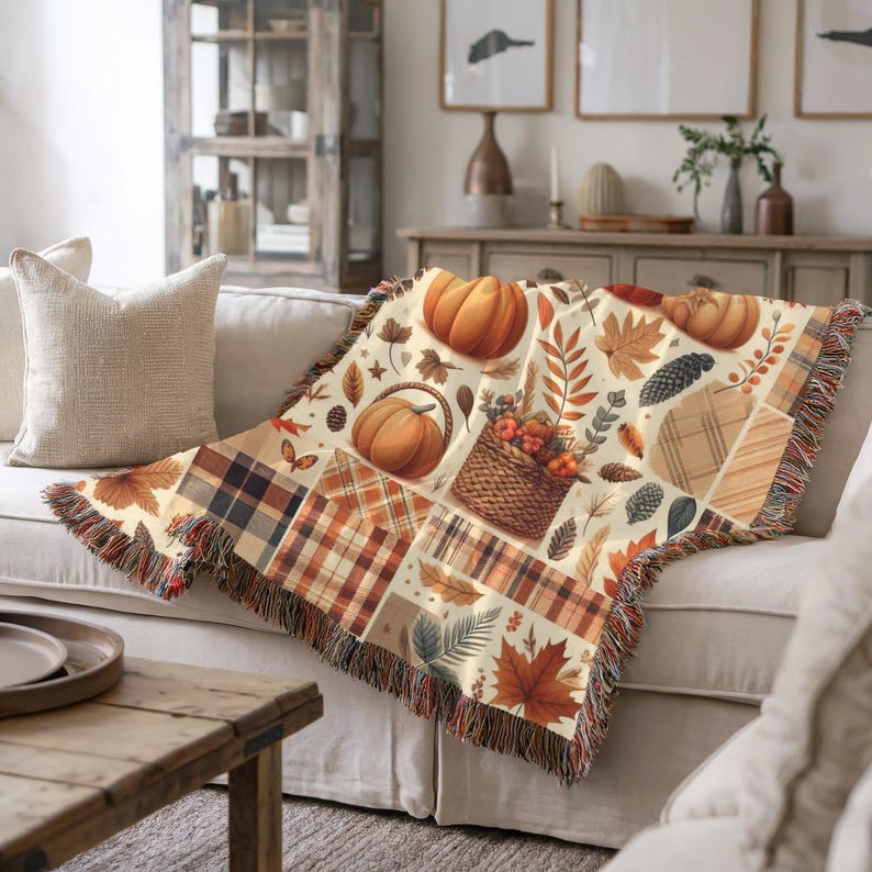 May include: A woven throw blanket featuring a patchwork design with autumn-themed illustrations. The blanket showcases pumpkins, fall leaves, and a basket of harvest goods in shades of orange, brown, and cream. The blanket has a fringed edge.