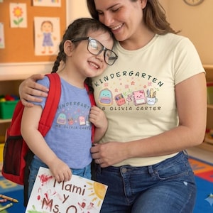 Puede incluir: Una camiseta azul claro y una camiseta color crema, ambas con un gráfico de útiles escolares con el texto KINDERGARTEN MS OLIVIA CARTER. El gráfico incluye una mochila, un estuche, una chincheta, un cuaderno y una taza con bolígrafos.