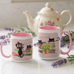 Puede incluir: Dos tazas de cerámica blanca con interior y asa de color rosa. Las tazas presentan ilustraciones de gatos negros sobre pilas de libros coloridos, una flor amarilla en maceta y el texto Olivia Carter y Read More Books impreso.