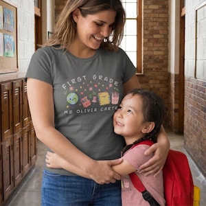 Puede incluir: Una camiseta gris oscuro de manga corta con el texto FIRST GRADE y MS OLIVIA CARTER junto a ilustraciones coloridas de un globo terráqueo, un estuche, una chincheta, un cuaderno y un portalápices rodeados de estrellas y flores.