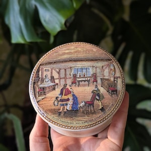 May include: A round, cream-colored decorative box with a detailed illustration of a room with people in period clothing. The box has text around the edge that reads "THE ROOM IN WHICH SHAKESPEARE WAS BORN 1564 STRATFORD ON AVON."