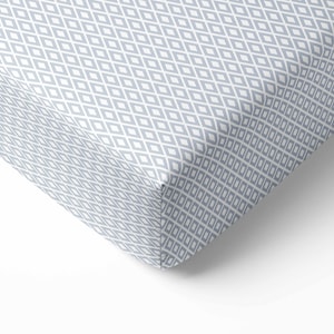 May include: A corner of a fitted sheet with a repeating diamond pattern in light gray and white. The fabric is folded over, showing the edge of the sheet against a white background. The sheet appears to be made of a soft material.