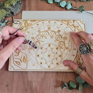 Op de afbeelding: Een houten divinatiebord met gegraveerde symbolen en een cirkelvormig ontwerp, met de woorden "Feu", "Eau", "Terre" en "Air". Een paarse en zilveren pendel wordt boven het bord gehouden. Het bord staat op een houten oppervlak.
