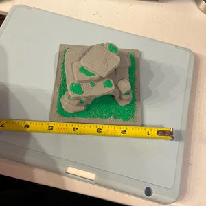 May include: A 3D-printed miniature terrain piece, featuring gray rock formations with green accents on a green base. The piece is approximately 4 inches long, as measured by a yellow tape measure. The terrain sits on a light blue surface.