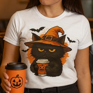 Puede incluir: Camiseta blanca con un gato negro con sombrero de bruja, sosteniendo una taza de café con un diseño de calabaza. El gato está rodeado de murciélagos y detalles en naranja. La persona sostiene una taza de café naranja con un diseño de calabaza.