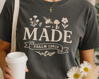 May include: Dark grey t-shirt with the word "MADE" in a cream font, decorated with floral and insect illustrations. The text "PSALM 139:14" is below "MADE". A gold cross necklace is visible. The person is holding a coffee cup, a book, and flowers.