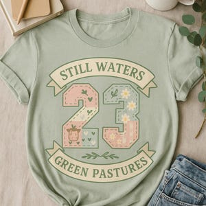 May include: A light green t-shirt with the text "Still Waters Green Pastures" in a banner design. The number "23" is in the center, decorated with floral and cow graphics. The shirt is folded and knotted at the bottom.