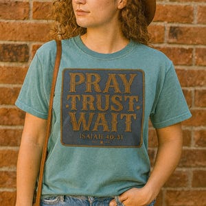 May include: Teal short-sleeve t-shirt with a rectangular graphic that reads "PRAY TRUST WAIT" in brown lettering, with "ISAIAH 40:31" below. The shirt is worn with blue jeans and a brown hat.