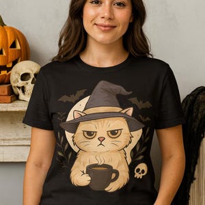 Könnte beinhalten: Schwarzes T-Shirt mit einer Cartoon-Katze, die einen Hexenhut trägt und eine Kaffeetasse hält. Das Design umfasst Fledermäuse, einen Totenkopf und einen Vollmond, perfekt für Halloween. Die Katze hat einen mürrischen Gesichtsausdruck.