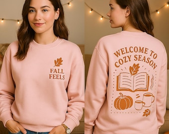 May include: A light pink sweatshirt with the text "FALL FEELS" on the front and "WELCOME TO COZY SEASON" on the back. The back design includes a pumpkin, book, and mugs in a warm brown color. The sweatshirt is a comfortable, casual style.