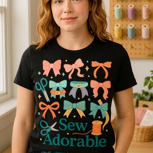 Puede incluir: Camiseta negra con un diseño de lazo colorido y el texto "Sew Adorable". El diseño incluye tijeras y una bobina de hilo. La modelo lleva vaqueros azules.