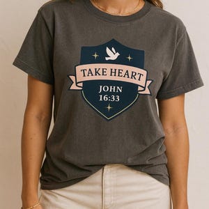 May include: Dark gray t-shirt featuring a shield design with the words "TAKE HEART" on a pink banner. Below, it reads "JOHN 16:33". The shield includes a white dove and stars. The shirt is a casual wear item.