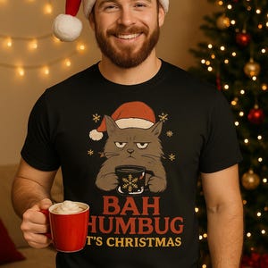 May include: Black t-shirt with a grumpy cat wearing a Santa hat, holding a coffee mug. The text reads "BAH HUMBUG, T'S CHRISTMAS". A person wearing a Santa hat holds a red mug. Christmas tree in the background.