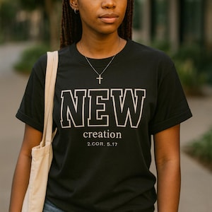 Op de afbeelding: Zwart T-shirt met het woord "NEW" in witte blokletters, gevolgd door "creation" en de Bijbeltekst 2.COR. 5.17. De persoon draagt een zilveren kruis ketting en een beige draagtas.