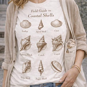 May include: Cream-colored t-shirt featuring a field guide to coastal shells, with illustrations of various seashells and their names. The shirt is worn with blue jeans and a beige cardigan. The text "Field Guide to Coastal Shells" is printed at the top.