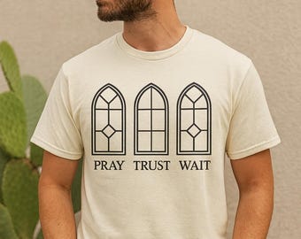 May include: Cream-colored t-shirt with a black graphic of three arched windows and the words "PRAY TRUST WAIT" printed below. The person in the image is wearing a brown corduroy cap and blue jeans.