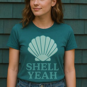 Op de afbeelding: Tealkleurig T-shirt met een licht-teal schelpgrafiek en de woorden "SHELL YEAH" in bijpassende teal kleur. Het shirt wordt gecombineerd met een blauwe jeans. De achtergrond is een teal-kleurige houten muur.
