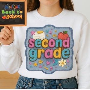 May include: White sweatshirt with a colorful "Second Grade" graphic. The design includes an apple, stars, and a flower. A small chalkboard with "Welcome Back to School" is held in the background.