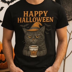 Puede incluir: Camiseta negra con el texto "HAPPY HALLOWEEN" en naranja. La camiseta presenta un gato negro gruñón con un sombrero de bruja y sosteniendo una taza de café con un diseño de calabaza. También se incluyen murciélagos.