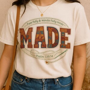 May include: Cream-colored t-shirt with the word "MADE" in a patchwork design, featuring brown, blue, and orange hues. The text "Fear fully & wonder fully made" arches above the word, with "Psalm 139:14" below. A needle and thread graphic is also present.