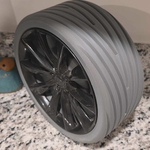May include: A gray and black car wheel with a unique ribbed tire design. The wheel has a dark, metallic finish with multiple spokes. The tire has a series of parallel grooves, creating a textured appearance. The wheel is set against a speckled countertop.