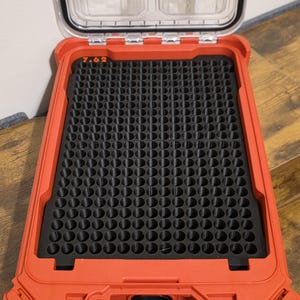 May include: A red storage case with a clear lid, open to reveal a black foam insert with many circular holes. The text "7.62" is printed inside. The case is rectangular and designed for storing small items. The case is likely used for organizing ammunition.