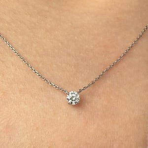 Peut inclure: Un collier &agrave; cha&icirc;ne fine de couleur argent&eacute;e dot&eacute; d'un pendentif unique en pierre claire ronde &agrave; taille brillant, fix&eacute; dans un sertissage simple &agrave; quatre griffes. Le collier repose sur le cou, pr&eacute;sentant un design &eacute;pur&eacute; avec une fine cha&icirc;ne en m&eacute;tal poli.