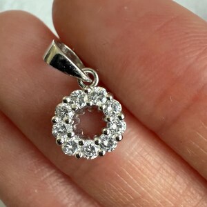 May include: A circular silver pendant featuring a cluster of ten small, clear, round-cut stones arranged in a ring formation. The pendant includes a polished silver bail at the top for attachment to a chain, showcasing a bright, reflective metallic finish.