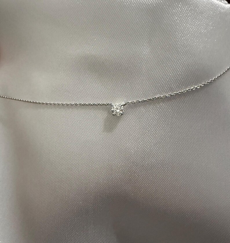 Peut inclure: Un collier &agrave; cha&icirc;ne fine de couleur argent&eacute;e orn&eacute; d'une unique petite pierre ronde transparente &agrave; facettes, fix&eacute;e au centre par un sertissage &agrave; griffes simple. La fine cha&icirc;ne m&eacute;tallique s'&eacute;tend horizontalement, pr&eacute;sentant un design &eacute;pur&eacute;.
