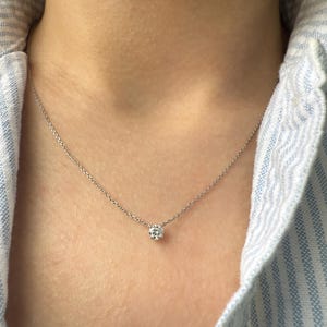Peut inclure: Un collier &agrave; cha&icirc;ne fine de couleur argent&eacute;e orn&eacute; d'une unique petite pierre pr&eacute;cieuse transparente, ronde et taill&eacute;e en brillant, fix&eacute;e dans un sertissage m&eacute;tallique simple &agrave; quatre griffes. Le collier repose sur le cou avec une cha&icirc;ne fine.