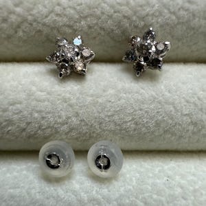 May include: A pair of silver-toned stud earrings, each featuring a flower design with multiple small, clear gemstones. The earrings are displayed with two clear earring backs on a soft, white surface.