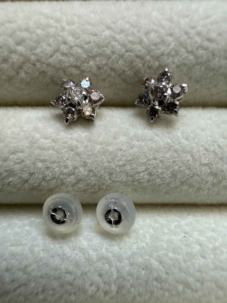 May include: A pair of silver-toned stud earrings, each featuring a flower design with multiple small, clear gemstones. The earrings are displayed with two clear earring backs on a soft, white surface.