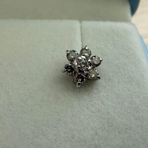 May include: A silver-toned, flower-shaped stud earring with multiple clear and dark-toned gemstones. The earring is set against a light blue-green background, suggesting it is in a jewelry box.