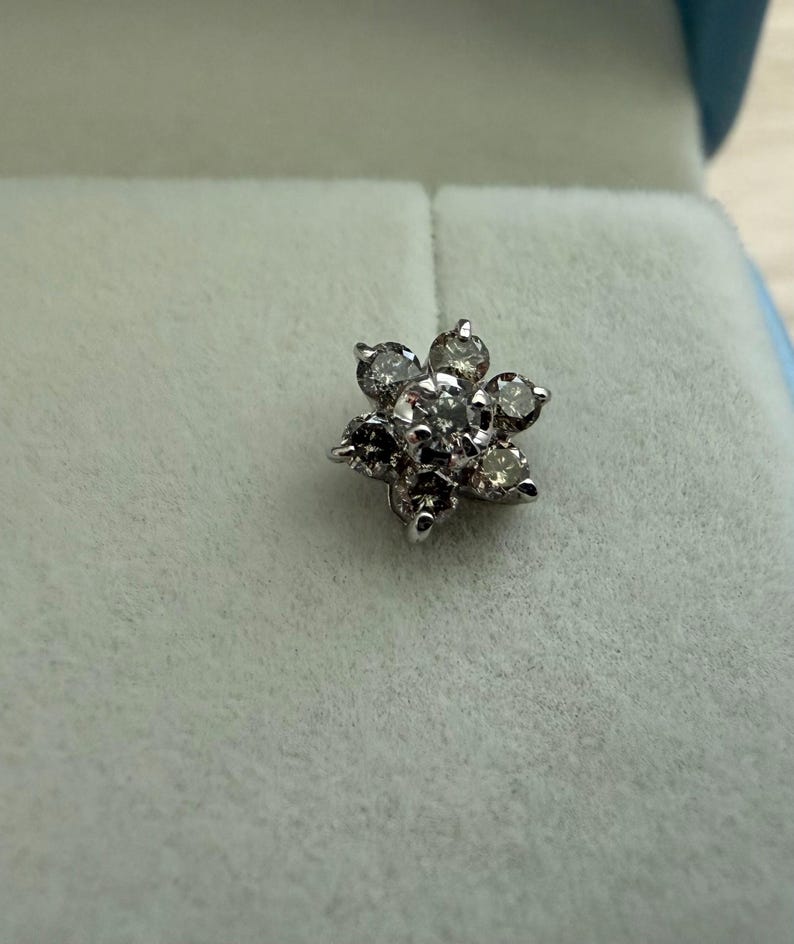 May include: A silver-toned, flower-shaped stud earring with multiple clear and dark-toned gemstones. The earring is set against a light blue-green background, suggesting it is in a jewelry box.