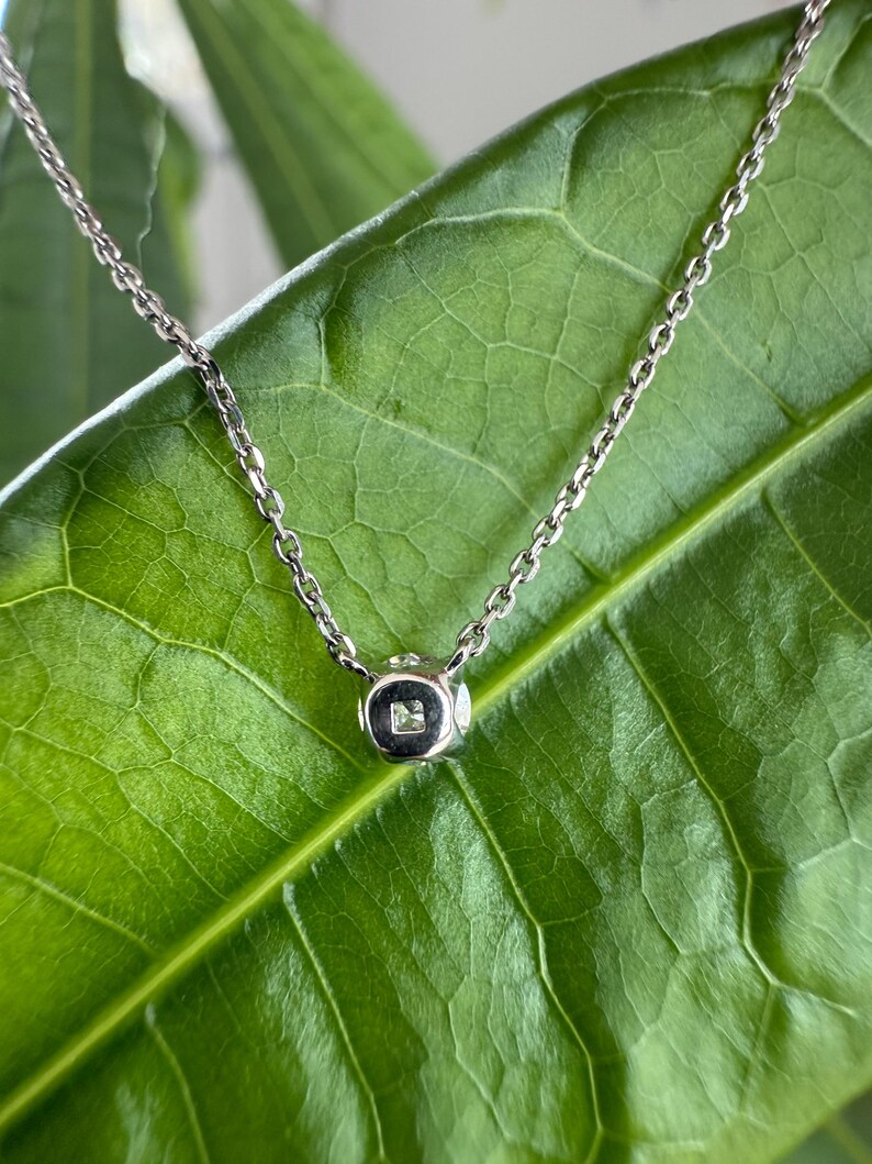 Peut inclure: Un collier &agrave; cha&icirc;ne en m&eacute;tal argent&eacute; dot&eacute; d'un petit pendentif en forme de cube. Le pendentif pr&eacute;sente une finition lisse et polie et contient une unique petite pierre claire de taille carr&eacute;e sertie en son centre, suspendu &agrave; une cha&icirc;ne fine.