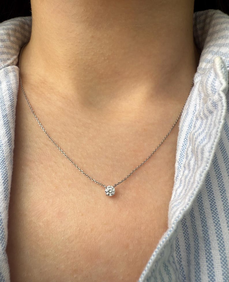 Peut inclure: Un collier &agrave; cha&icirc;ne fine de couleur argent&eacute;e dot&eacute; d'un pendentif unique en pierre claire ronde &agrave; taille brillant, fix&eacute; dans un sertissage simple &agrave; quatre griffes. Le collier repose sur le cou, pr&eacute;sentant un design &eacute;pur&eacute; avec une fine cha&icirc;ne en m&eacute;tal poli.