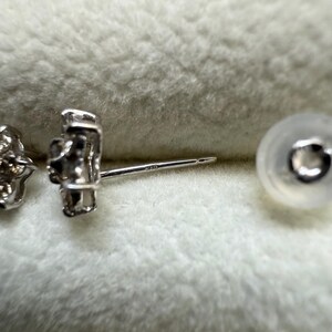 May include: Close-up of a silver stud earring with a flower-like design. The earring post is visible, along with a clear earring back. The earring is set against a soft, textured background.