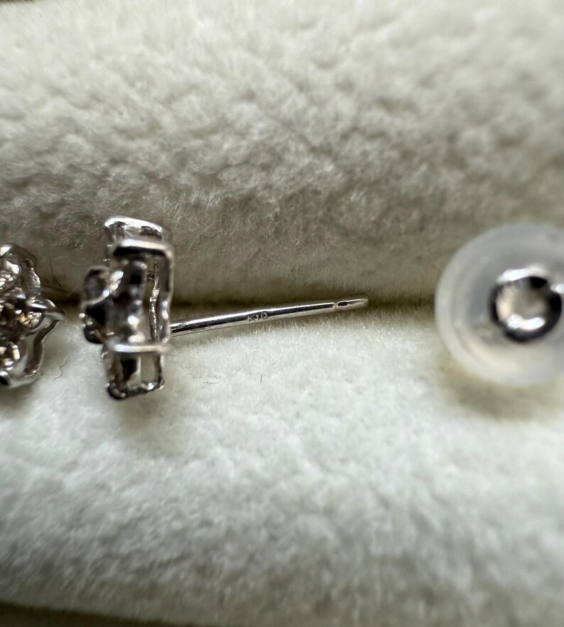 May include: Close-up of a silver stud earring with a flower-like design. The earring post is visible, along with a clear earring back. The earring is set against a soft, textured background.