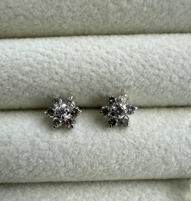 May include: A pair of flower-shaped stud earrings. Each earring features a central clear stone surrounded by six smaller, darker stones, creating a floral design. The earrings are set against a soft, light gray background.