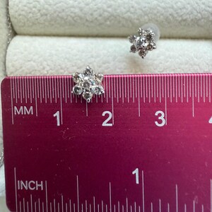 May include: A pair of silver-toned stud earrings in a flower design. Each earring features multiple clear gemstones. The earrings are approximately 0.5 inches in diameter. The earrings are displayed on a pink ruler.