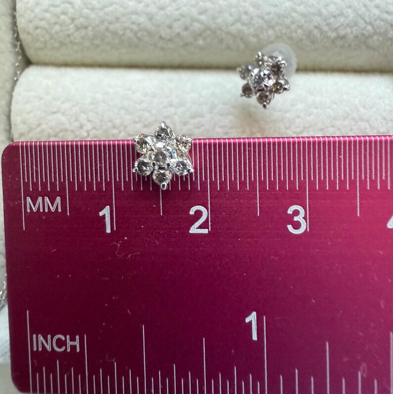May include: A pair of silver-toned stud earrings in a flower design. Each earring features multiple clear gemstones. The earrings are approximately 0.5 inches in diameter. The earrings are displayed on a pink ruler.