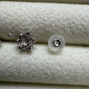 May include: A close-up of a silver-colored floral stud earring with a clear earring back. The earring has a small, round, clear stone in the center. The earring back is a clear, rounded disc with a metal insert.