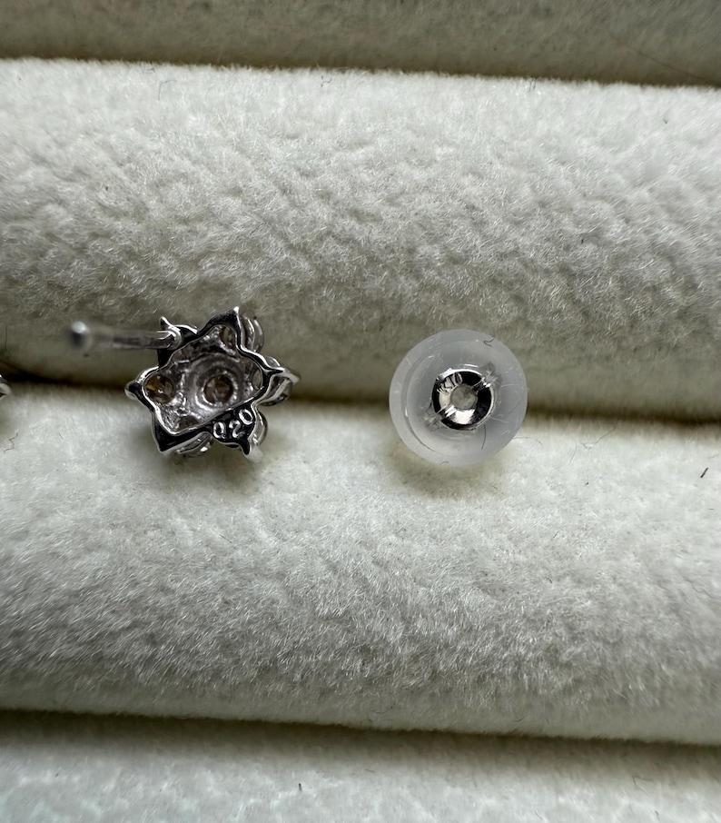 May include: A close-up of a silver-colored floral stud earring with a clear earring back. The earring has a small, round, clear stone in the center. The earring back is a clear, rounded disc with a metal insert.