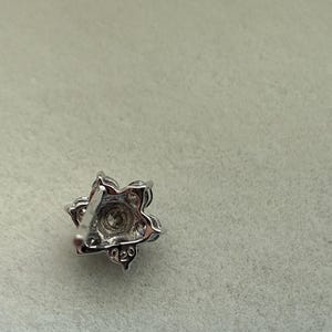 May include: A silver-toned, floral-shaped stud earring with a central clear stone and surrounding smaller stones. The earring has a post backing and is set against a light gray background. The earring is marked with "020".