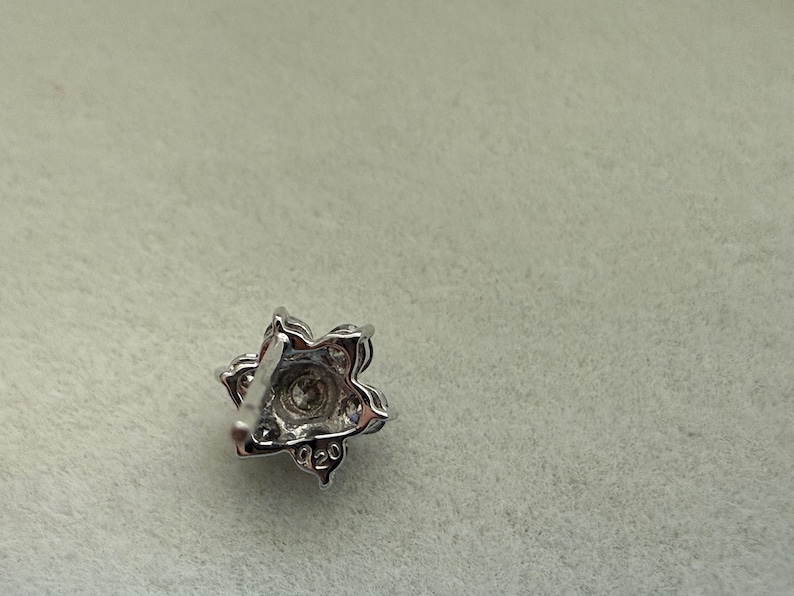 May include: A silver-toned, floral-shaped stud earring with a central clear stone and surrounding smaller stones. The earring has a post backing and is set against a light gray background. The earring is marked with "020".