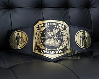 Puede incluir: Un cinturón negro de campeón de ortografía con detalles dorados. La placa central presenta una abeja, notas musicales y un libro abierto. El cinturón tiene dos placas laterales con figuras de personas. Se muestra el texto "SPELLING BEE CHAMPION".