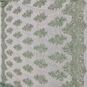 May include: White fabric with a delicate, all-over pattern of light green floral embroidery. The edge features a scalloped border with intricate beadwork, creating a luxurious and elegant textile.
