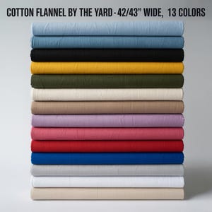 Könnte beinhalten: Ein Stapel gerollter Baumwollflanellstoffe in verschiedenen Farben, darunter Blau, Schwarz, Gelb und Rot. Der Text oben lautet "COTTON FLANNEL BY THE YARD - 42/43" WIDE, 13 COLORS". Der Stoff ist 106,68 bis 109,22 cm breit.